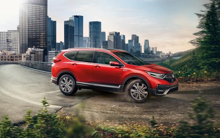 Passenger-side view of the 2022 CR-V Hybrid Touring in Radiant Red Metallic, driving from a city into a forest.