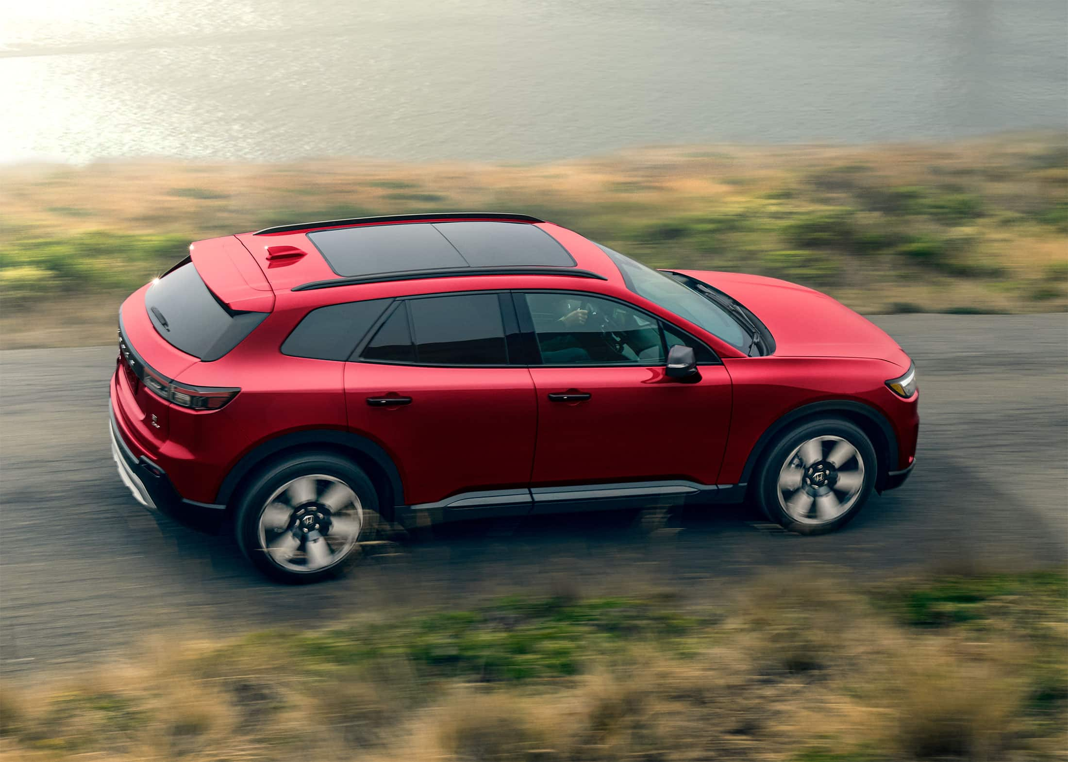 Vista de 7/8 desde el lado del pasajero trasero de la Honda Prologue Elite 2024 con tracción integral en Scarlett Red Metallic, que se muestra conduciendo con un puente grande de fondo.